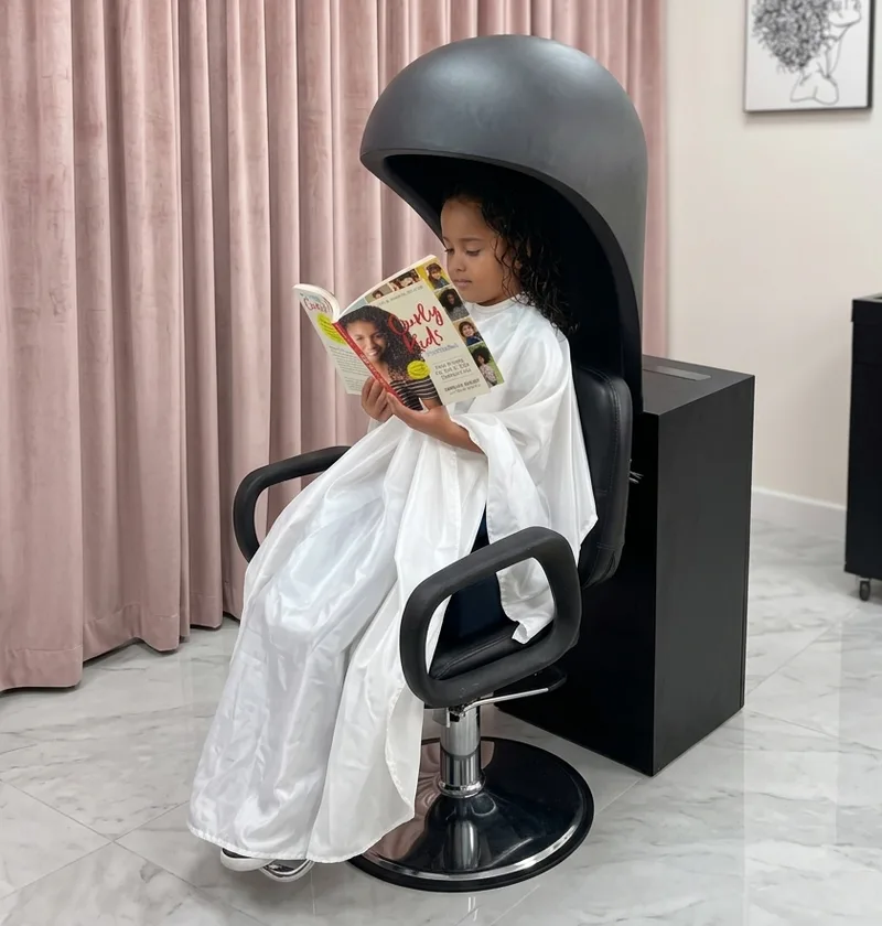A young client with curly hair reading a book in the salon chair at A Curl’s Best Friend, Lake Forest, CA
